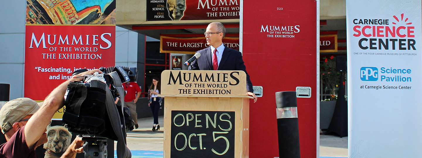 At the entrance to the Science Center, Science Center Director speaks behind a podium to the visiting reporters