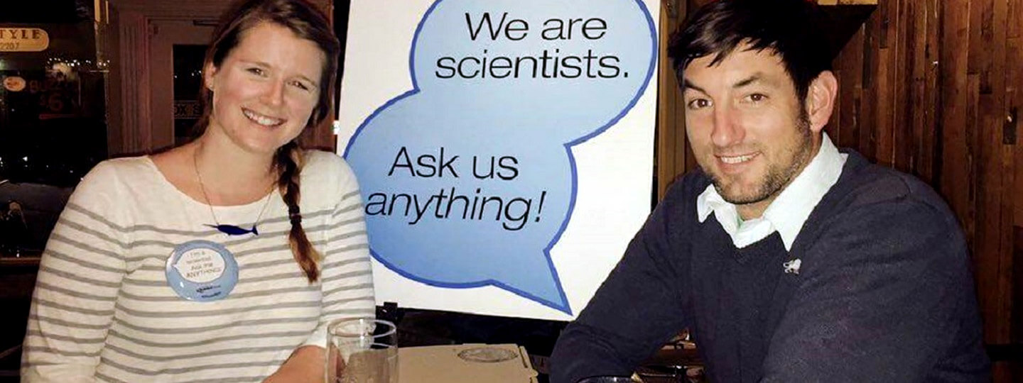 A female and male scientists sitting at a table with a sign saying we are scientists ask us anything