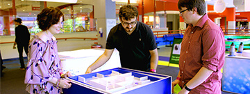 Two men and one woman engaging with a large blue square exhibit with moveable white pieces inside